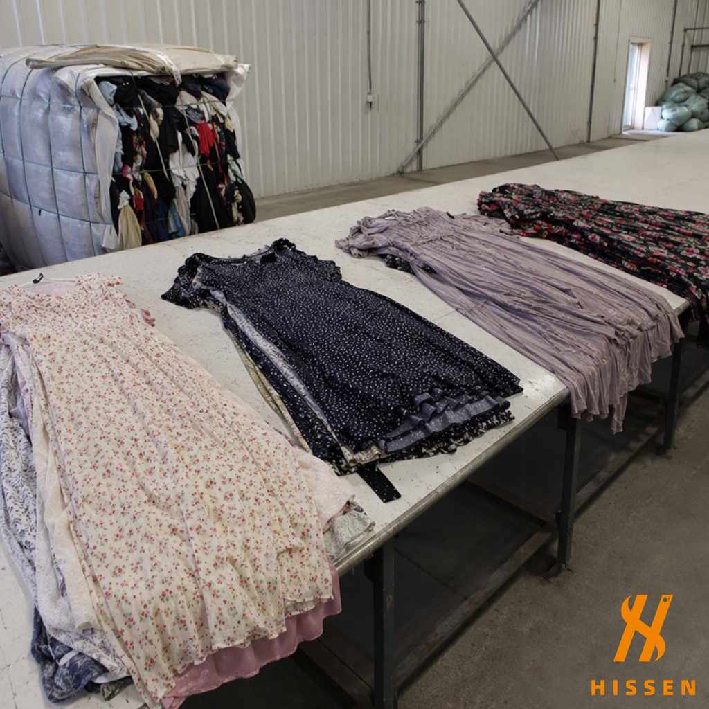 Four groups of used women's dresses laid out separately on a sorting table with an open clothing bale behind