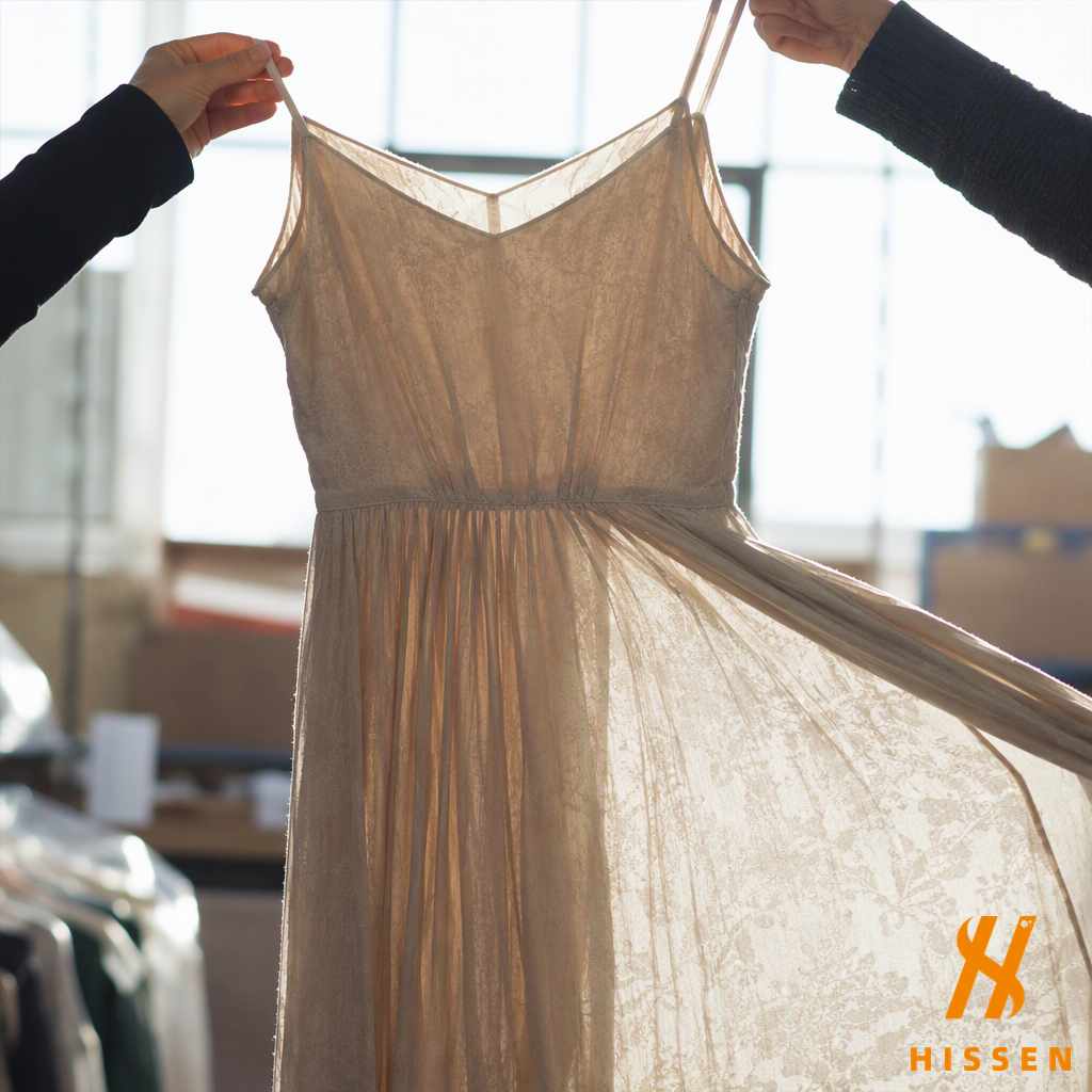 Hands holding a pale unlined long dress against a bright window, daylight showing the fabric is sheer