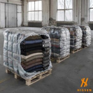 Compressed bales of used winter clothing on pallets in a warehouse, the nearest opened to show folded wool coats and padded jackets
